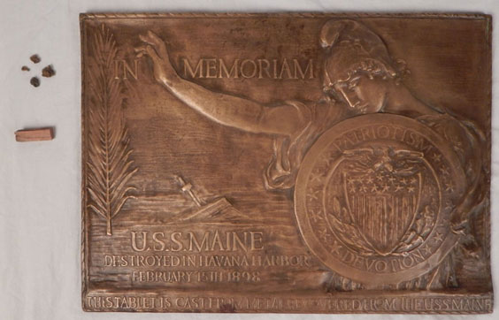 A memorial plaque for the U.S.S. Maine. There is an image of a person holding a shield. To the left side of the plaque are a few small pieces.