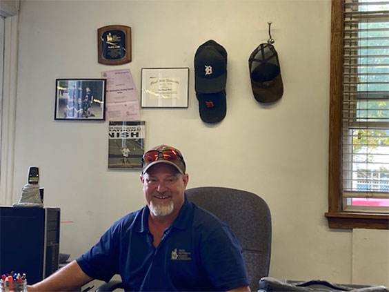 A man in a blue polo shirt and baseball cap sits behind a desk