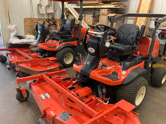 Two orange riding lawn mowers sit in a storage area