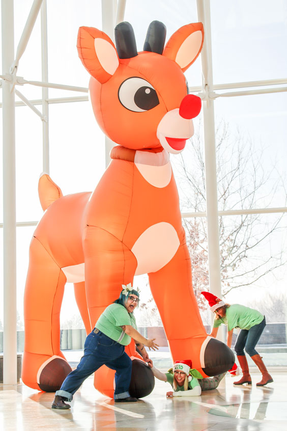 A giant inflatable reindeer with a red nose is stepping on a woman who is trying to be pulled out by the hand by a woman and by the feet by another woman