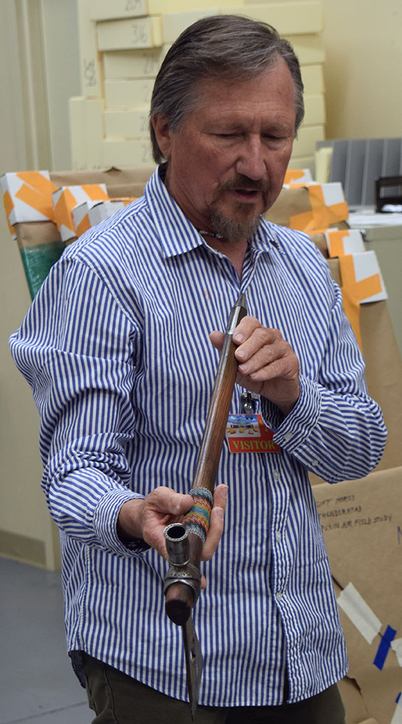 A dark haired man with a goatee who is wearing a blue and white pinstripe shirt stands holding an axe that doubles as a tobacco pipe