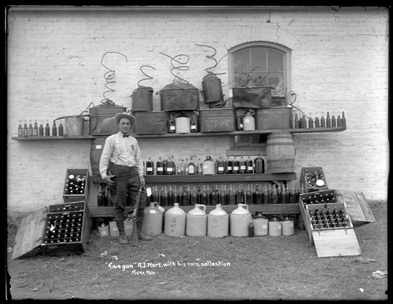 A man wearing a button up shirt, suspenders, bowtie, hat, slacks, and boots stands holding a gun in one hand while the other end of the gun is on the ground between his feet. Behind and around him are many bottles and jugs.