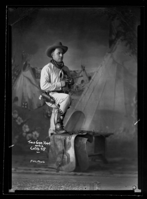 A young man in a cowboy hat, neck bandana, boots, and long sleeves and pants stands holding a hand gun with one leg perched on a bench. There is a gun holster around his belt. Behind him is a backdrop with at least three tipis and some flowers.