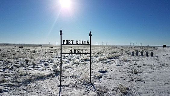 I metal sign reading Fort Dilts 1864 stands outsinde among frosted ground with windmills in the background. There are also 5 gravestones on the right side of the image in the middleground.