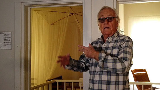 An older man with wihte hair and dark glasses wearing a black and white plaid shirt stands indoor in a house with two open doorways behind him.