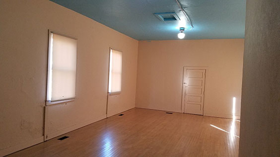An empty room with peach colored walls, a bluish gray ceiling, wood floors, one light on the ceiling, two windows with white shades pulled down, and a door on the adjacent wall that is the same color of the walls 