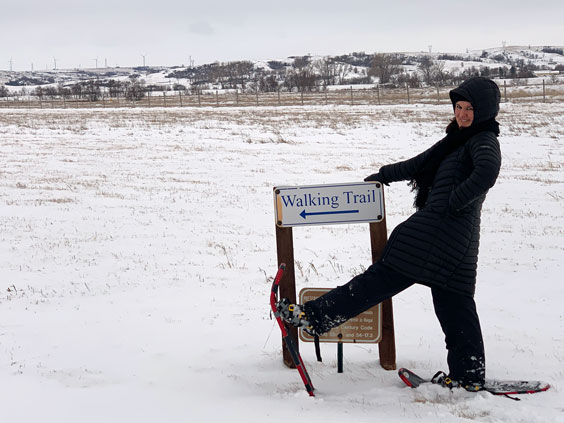 A woman dressed in black snow gear and red snowshoes stands with one leg up next to a sign that reads Walking Trail