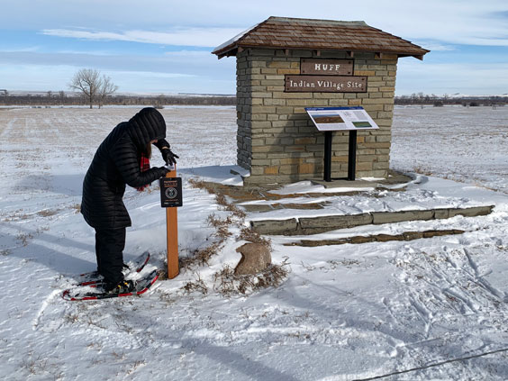 A woman dressed in black winter gear and red snowshoes stands at a stamping station next to a small brick structure reading Huff Indian Village Site with an interpretive panel in front of it