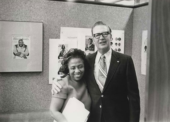 A white man in a dark suit, white button up shirt, and diagonal striped tie who is wearing dark rimmed glasses has his arm around a black woman wearing a short sleeved dress who is holding folders. Behind them are posters hanging on a wall.