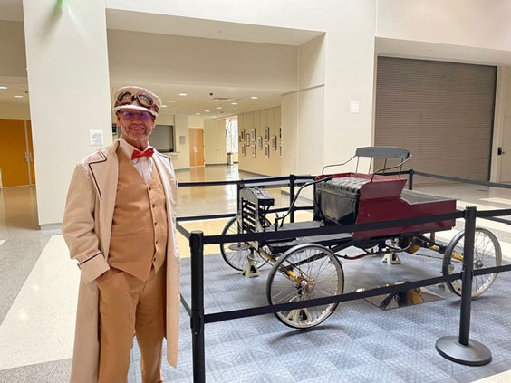 A gentleman with a short, white beard is wearing tan suit pants, vest, hat, and long overcoat, white button up shirt, orange bowtie, and old driving goggles on his hat. He is standing indoors next to a very old automobile that is on exhibit behind stanchions.