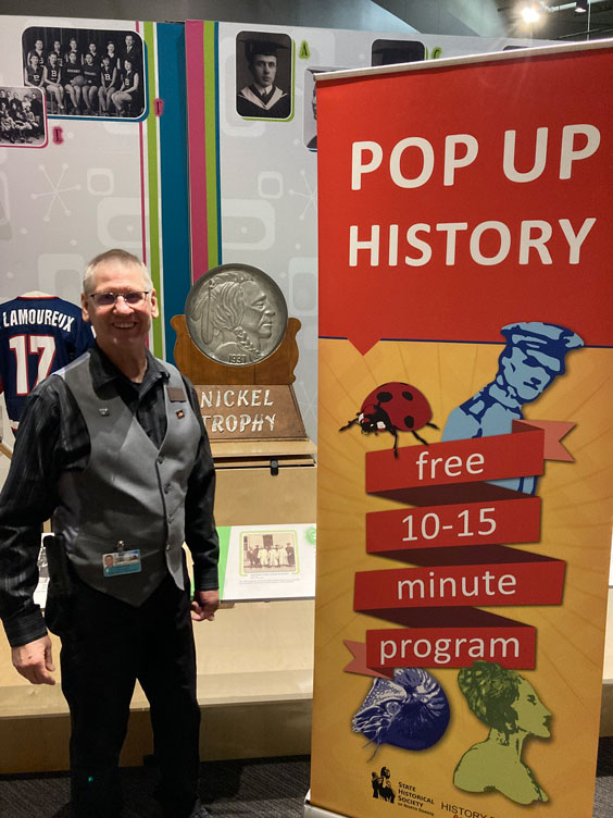 An older gentleman wearing black pants and button up shirt with a gray vest and dark rimmed glasses stands next to an orange and yellow pull-up banner that reads Pop Up History: free 10-15 minute program.