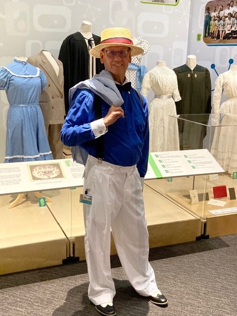An older gentleman is standing in front of a clothing exhibit wearing a straw hat with red ribbon around it, royal blue button up shirt with white cuffs and collar, black bowtie, black suspenders, white pants, and white and black shoes. He also has a white and blue pinstripe jacket over his shoulder.