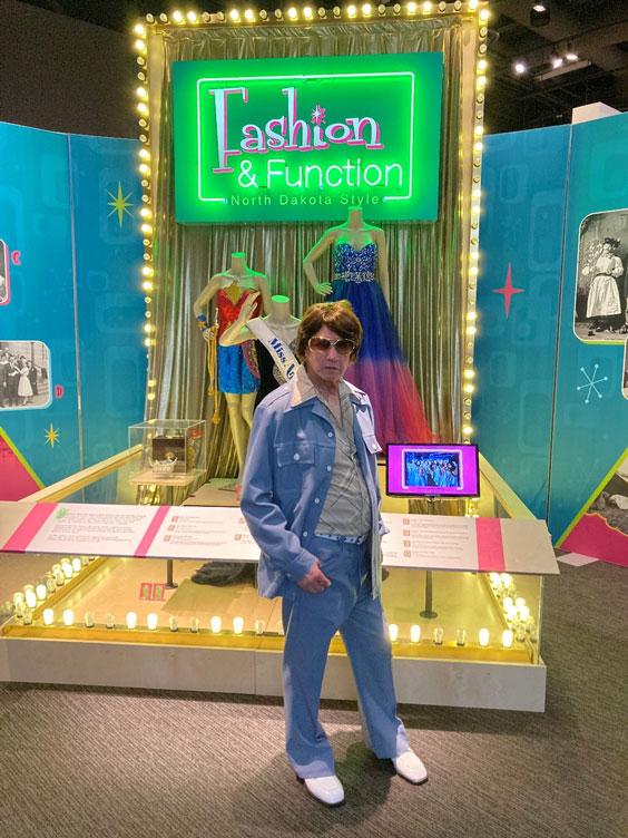 An older gentleman is standing in front of a Miss America exhibit with Fashion & Function North Dakota Style green and pink neon sign above it. He is wearing denim colored pants and button up jacket over a tan shirt, white shoes, and aviator type sunglasses.