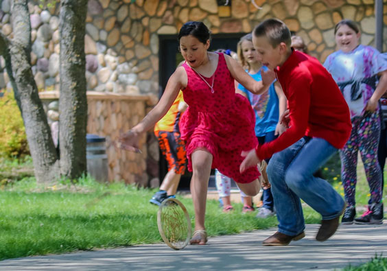 A young girl and boy are outdoors chasing after a hoop rolling on the ground. The girl is wearing a red dress, and the boy is wearing a red long sleeved shirt, blue jesans, and brown boots. There are other children watching in the background. 
