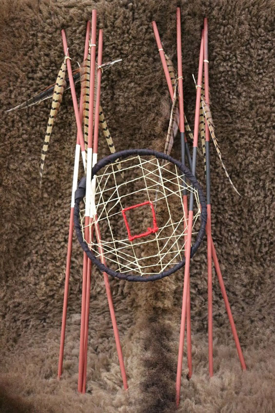 A brown hoop with light colored thread making a spider web like pattern in the inside with the middle being a red outlined square. To the sides of the hoop are pink sticks with a feather tied to the top. The ones on the left have a large white area towards the middle, and the ones on the right have a large black area towards the middle.