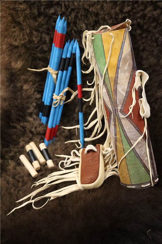 Many blue pointed sticks with a red and black band on each sit next to a carrying bag that is yellow, green, red, and white with gray separating the triangular colors. There are also four smaller sticks that are light tan in color with two of them having a black band across the middle. The carrying case for those is brown with tan on the sides.