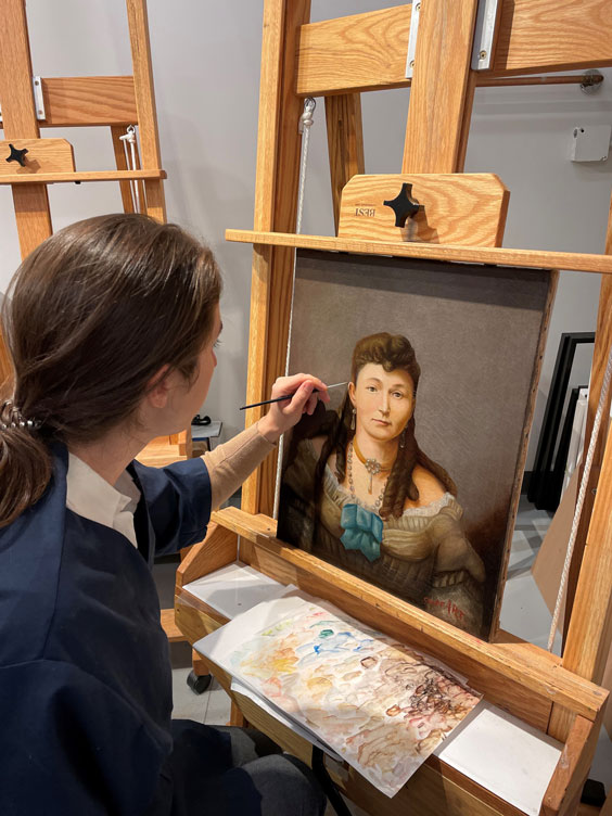 A woman with long, dark hair bulled back in a low ponytail wearing a dark blue shirt with a white shirt under it is touching up a painting of woman's portrait.