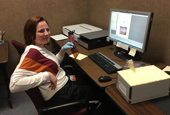 A young woman sits at a desk leaning back in the char. On the desk are a computer monitor, keyboard, mouse, microfilm scanner, and a couple other scanners.