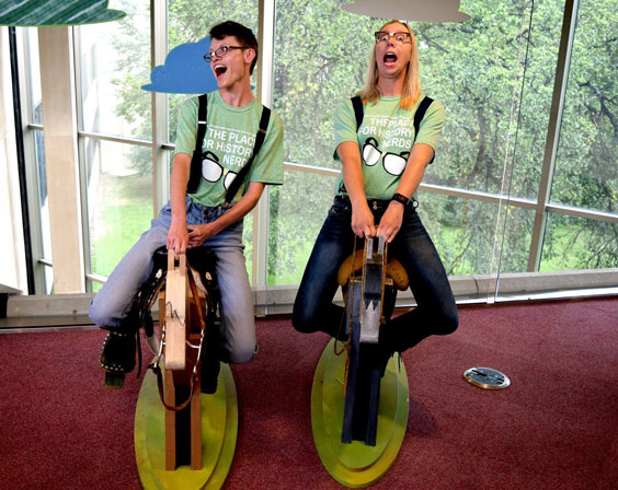 Two women dressed as nerds sit on wooden horses and pretend to ride them