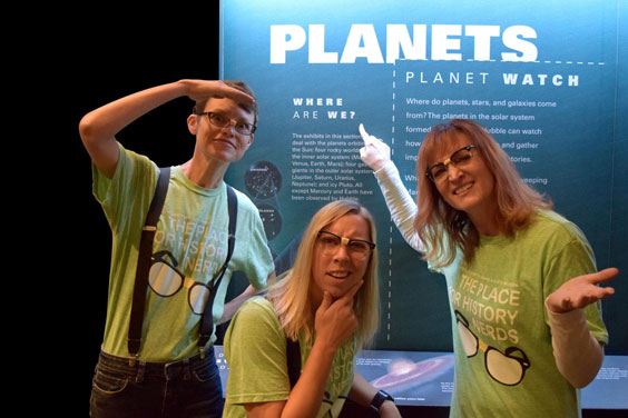 Three women dressed as nerds stand in front of a sign and are pointing to Where Are We