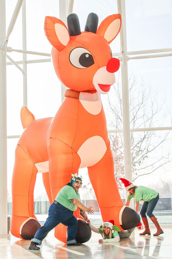 A woman dressed in a Santa hat and Santa slippers is getting stepped on by a large inflatable Rudolph the Red-Nosed Reindeer while two others dressed in Christmas attire try to pull her out