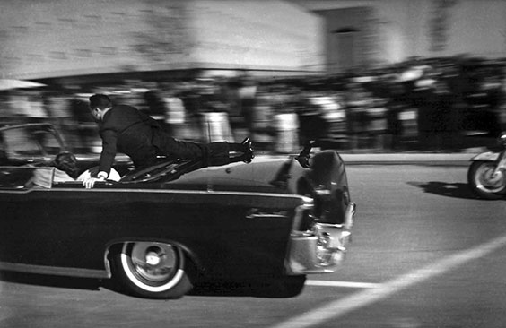 a man is shown partially in a convertible with one leg hanging across the back while he protects the passengers as the convertible drives off to safety