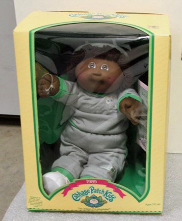 A black Cabbage Patch Kid doll with gray clothes and green trim sits in its yellow and green box.