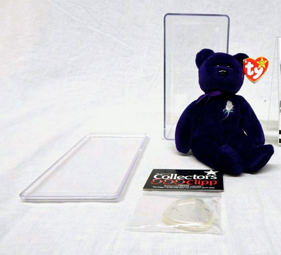 A purple Princess Diana TY Beanie Baby with a white rose on its left chest area. A plexiglass container sits behind it and collectors clipp for the tag sits in front of it.