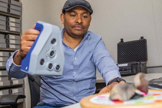 a man wearing a blue and white pinstripe shirt and a gray hat sits at a desk holding a #d scanner that looks similar to an iron but with 5 lenses on the underside.