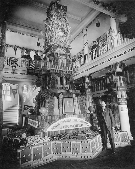 A man wearing a full suit stands in front of a corn display that says NORTH DAKOTA ENLIGHTNING THE WORLD. STATE OF OPPORTUNITY. Two men and three women stand in the balcony above..