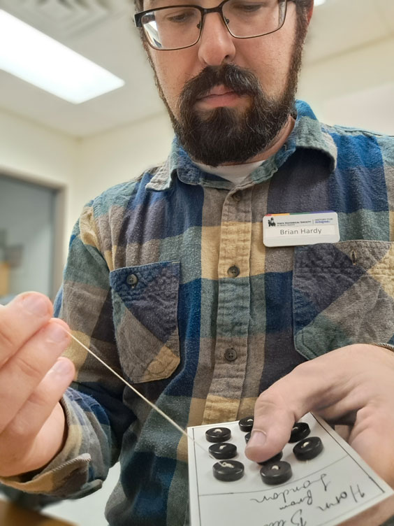 A man in a blue and tan plaid button up shirt with dark facial hair and glasses is sewing buttons onto a piece of paper