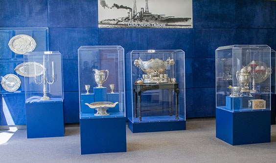 An exhibit display of USS North Dakota Silver Service items. The case bases and wall behind are blue, and an image of the USS North Dakota ship is on the wall behind