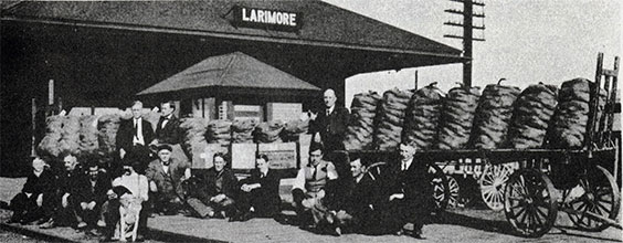 A group of people sit outside a building that reads Larimore. There are wagons with potato sacks on them.