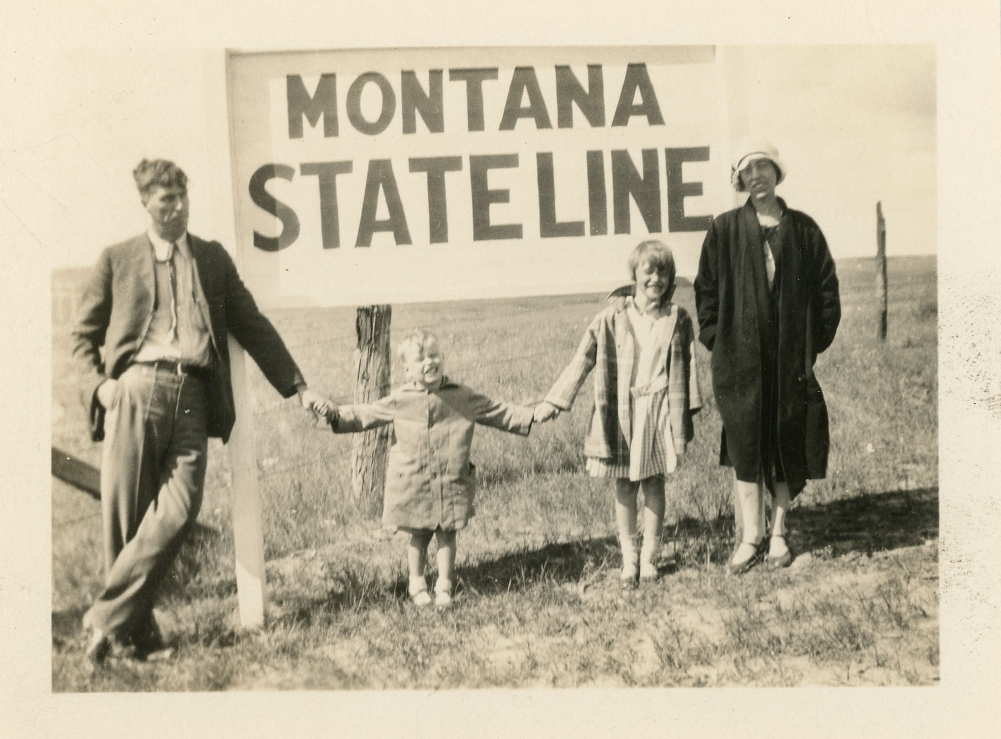 A family of 4 with the 2 children being toddler and elementary age stand outdoors in front of a sign that says Montana State Line. The photo was taken around 1930.
