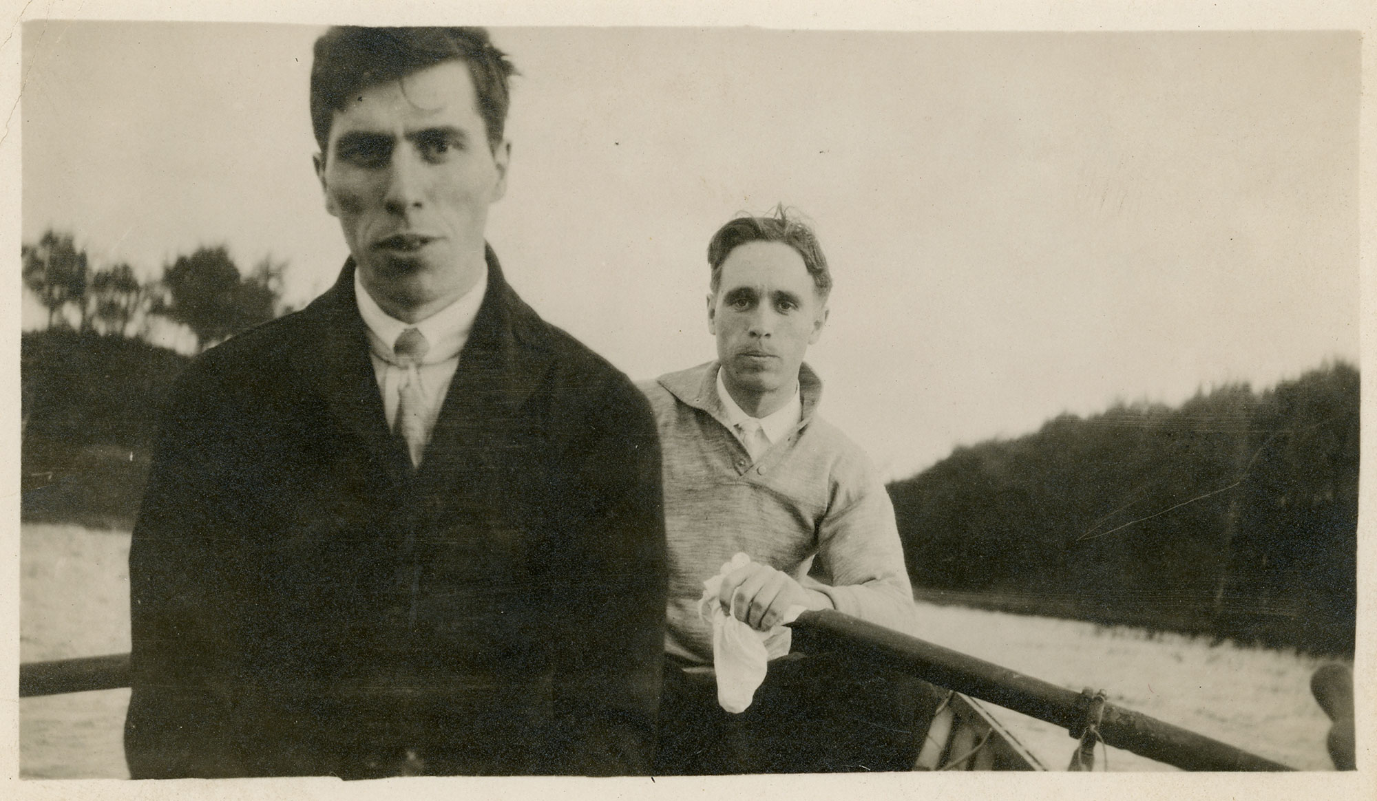 Two men in dress shirts and ties sit in a rowboat. The front man has dark hair and a dark jacket on. The back man who is rowing has lighter hair and a light colored collared pullover with a few buttons at the top.