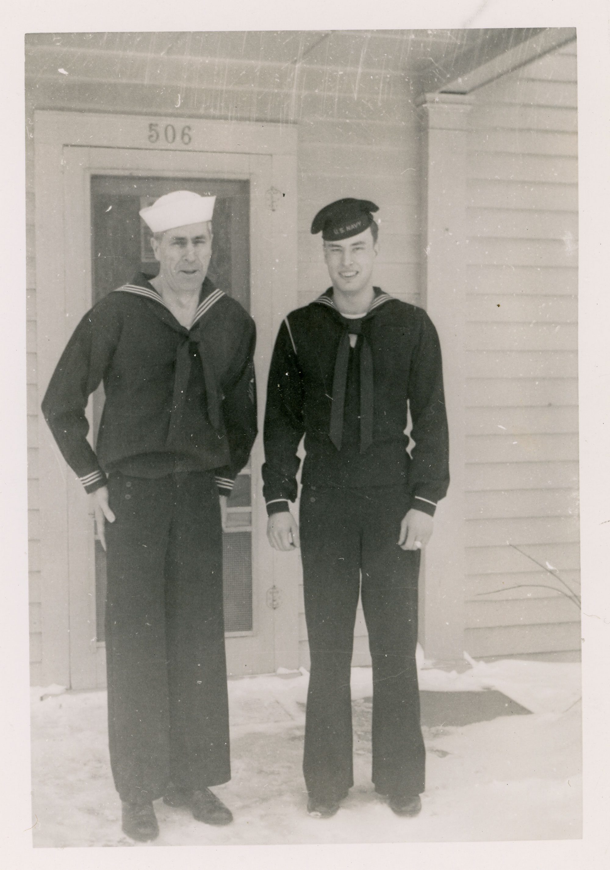 Two men in Navy uniforms stand outside a house with the number 506 above the door. The man on the left is middle-aged and has a white hat. The man on the right is younger and has a dark colored hat.