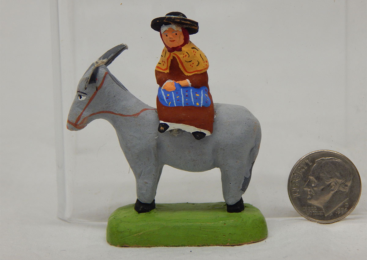 A figurine of an older lady with a large hat, red dress, yellow shawl over her shoulders and a blue blanket or apron on her lap sitting atop a gray donkey.