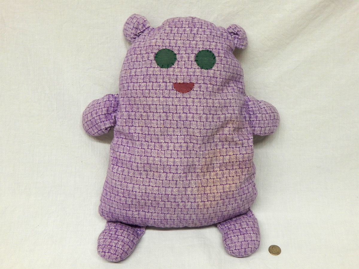 A purple stuffed animal that's sort of gumdrop shaped with little ears, arms, and legs and has black eyes and a red half circle shaped mouth.