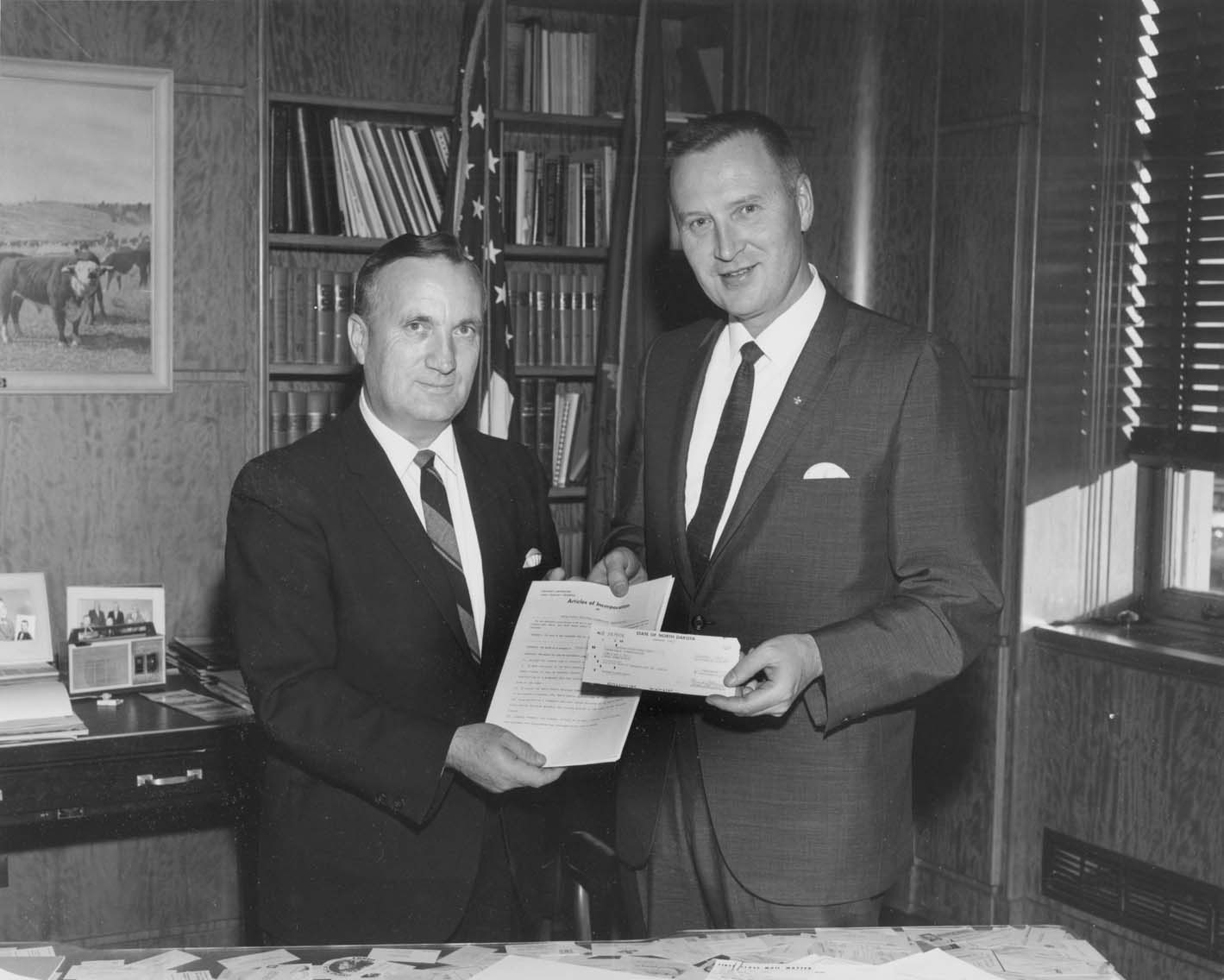 two men in suits and ties stand in an office holding two pieces of paper