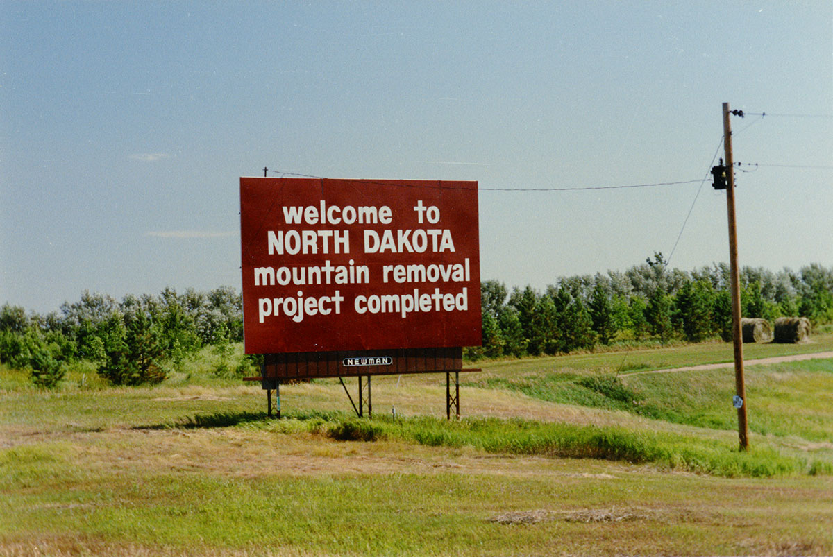 A brownish red billboard with white text that reads welcome to NORTH DAKOTA - mountain removal project completed