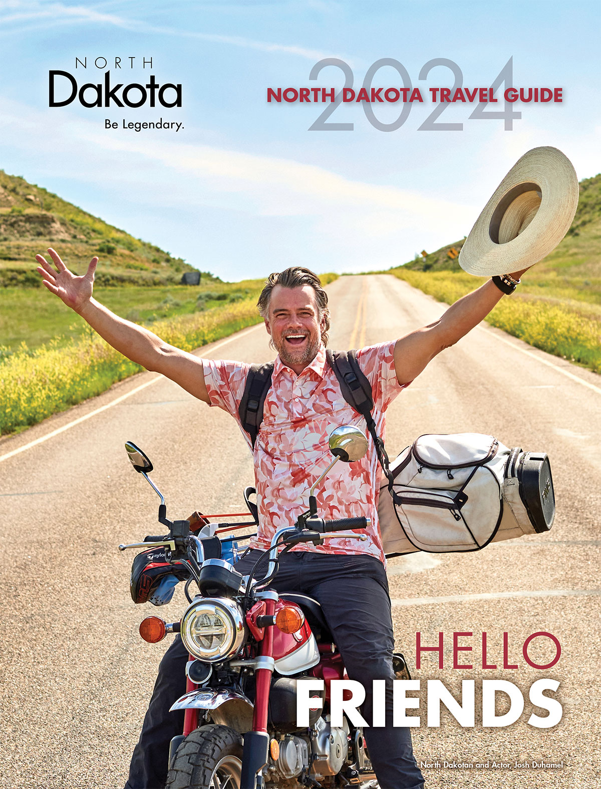 2024 North Dakota Travel Guide cover. A man holding a straw hat with a pink, red, and white floral short sleeved polo and dark bants sits on a red and white motorcycle with a set of golf clubs on his back. He is shown with his hands in the air on an open road with grassy hills to the sides of him.
