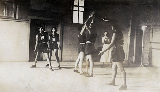 Five women play basketball while a woman stands in the background coaching or reffing