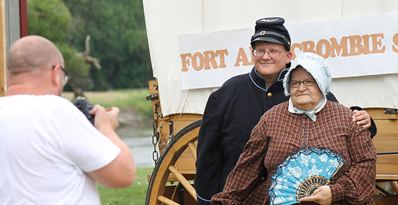 Two adults posing in old time clothing while a man takes their picture