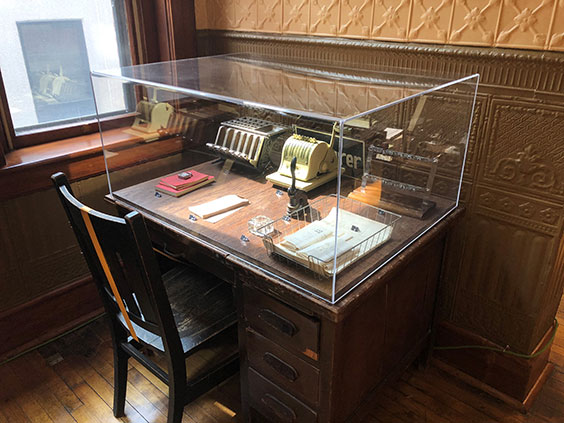 A glass or plastic case is sitting atop a desk with books, papers, and old desk gadgets, possibly adding machines, in it. There is a chair sitting in front of the desk.