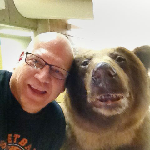 An adult man who is bald and wearing black glasses poses next to a bear