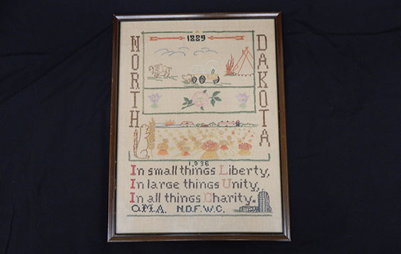 Framed beaded piece that says North Dakota 1889 - 1936 In small things Liberty, In large things Unity, In all things Charity. There are clouds, a bison, covered wagin, tipi, squirrel holding wheat, farmstead, and the state capitol depicted.