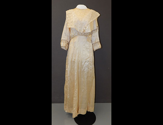 An off white/tan wedding dress. It is full length and has long sleeves. There is a draped part over the chest. Beaded fringe hands off of part of the chest drape and the sleeves.