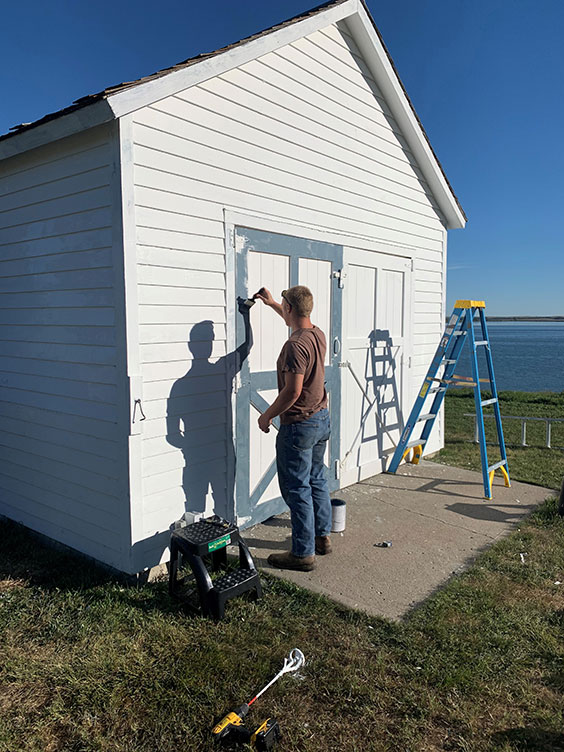 A man stands outside painting the trim of a door blue. The rest of the door and the building is white.