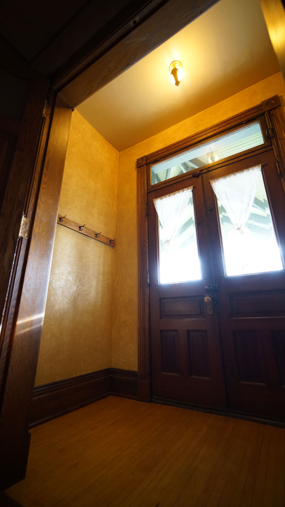 Entryway of a house with yellow wallpaper, dark wood trim, double dark wood doors with a large window in each and a windown spanning the top of both doors. There are also four coat hangers on one of the walls.