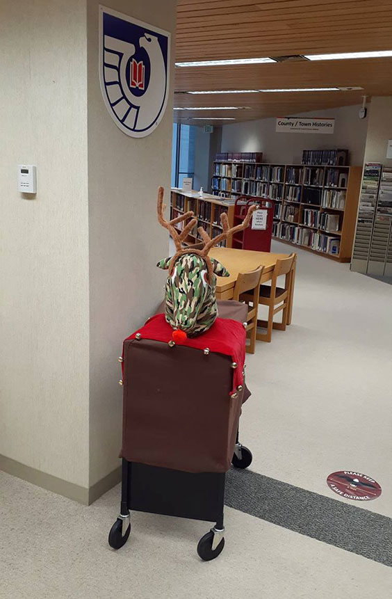 A black cart on wheels is shown with a brown cover over it and a reindeer head sitting atop it with a red draped cloth with bells to look like a reindeer.